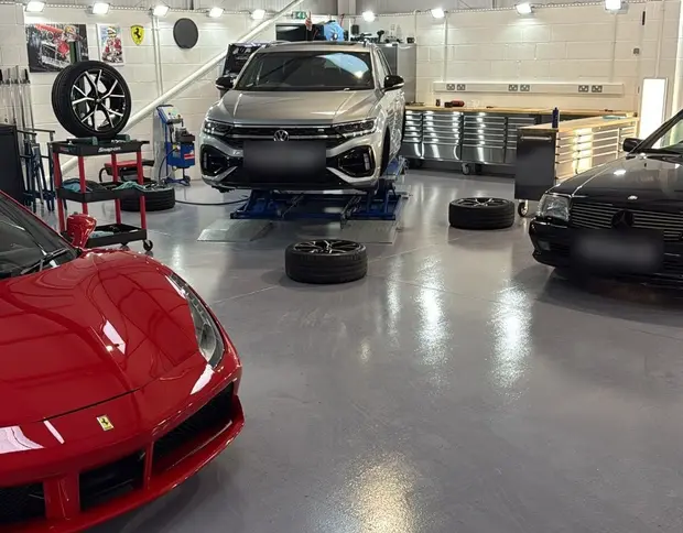 Three cars in the expanded ALC Elite workshop: a red Ferrari in the foreground, a silver SUV on a ramp and a black Mercedes