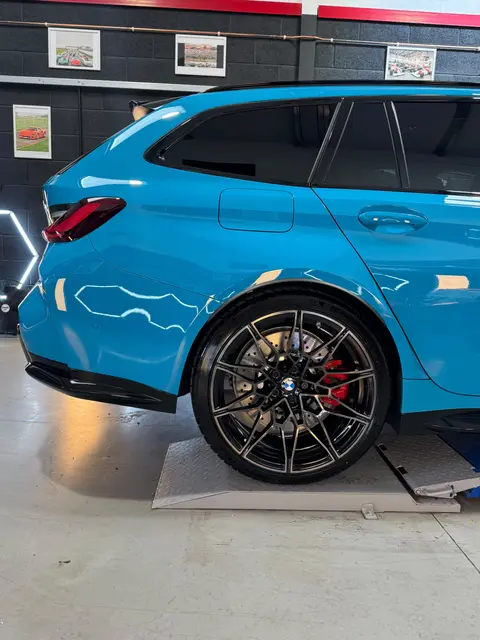 The side of a blue BMW M3 Touring on a ramp, with a black multi-spoke wheel and red brake caliper