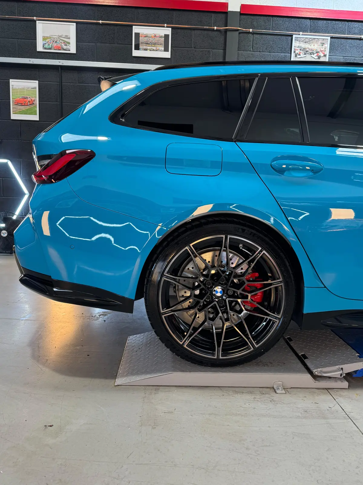 The side of a blue BMW M3 Touring on a ramp, with a black multi-spoke wheel and red brake caliper